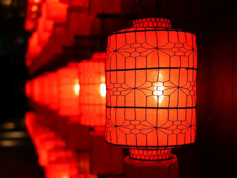 Uma fileira de lanternas chinesas vermelhas penduradas sobre uma rua, a mais próxima mostra sua estrutura de treliça e o brilho quente da lâmpada