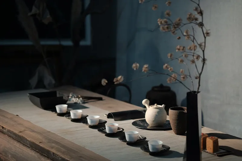 A small white porcelain gaiwan with tea leaves and two filled tasting cups on a dark wooden tea tray