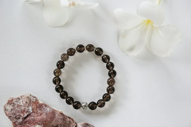 A strand of dark brown translucent crystal beads with silver spacers and a carved metal bead, laid on a white surface beside white flowers