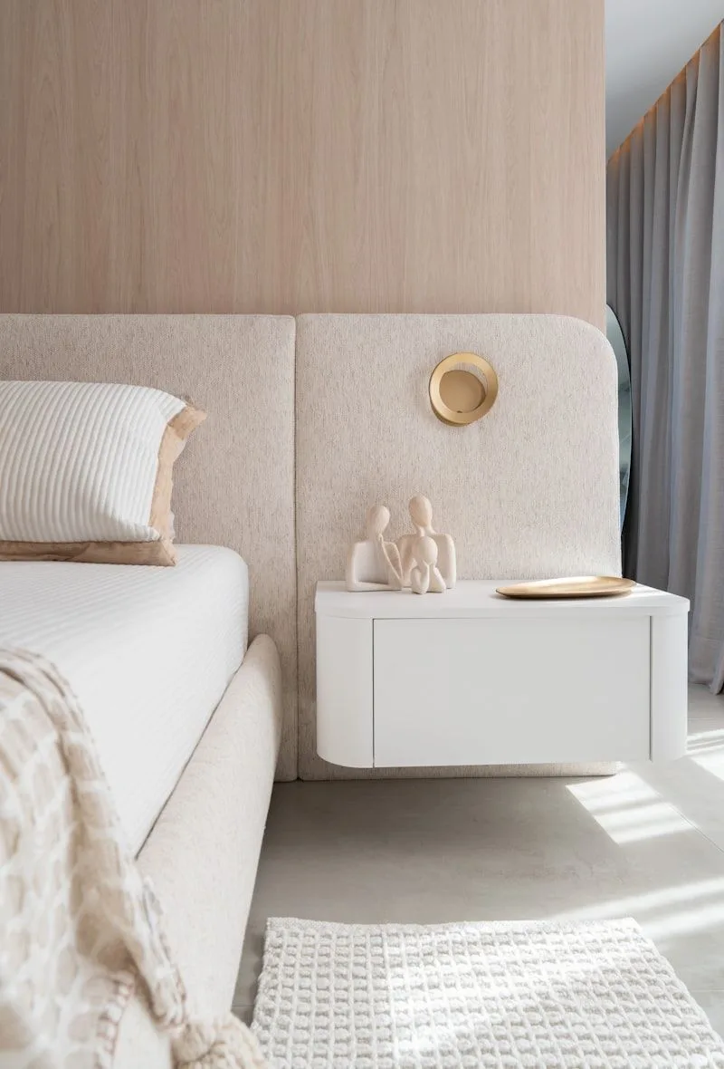 A clean minimalist bedroom with natural light, beige walls, and a wooden bed frame centered on the wall