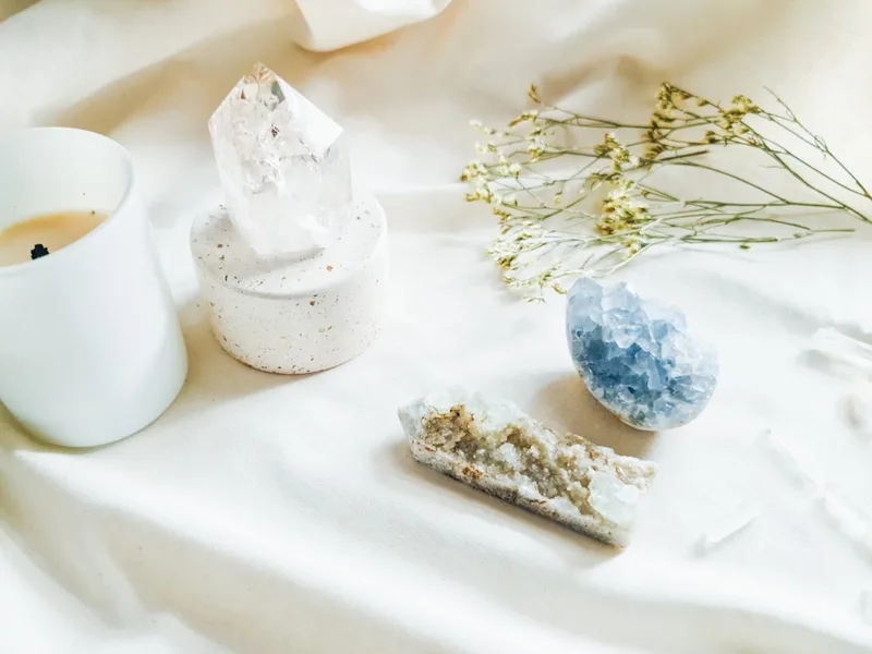 A lit white candle beside a clear quartz cluster and blue fluorite on cream fabric with dried flowers