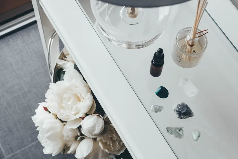 Uma coleção de cristais naturais em tons de branco, roxo e verde ao lado de um conta-gotas de óleo e peônias brancas sobre uma mesa de vidro, luz natural suave