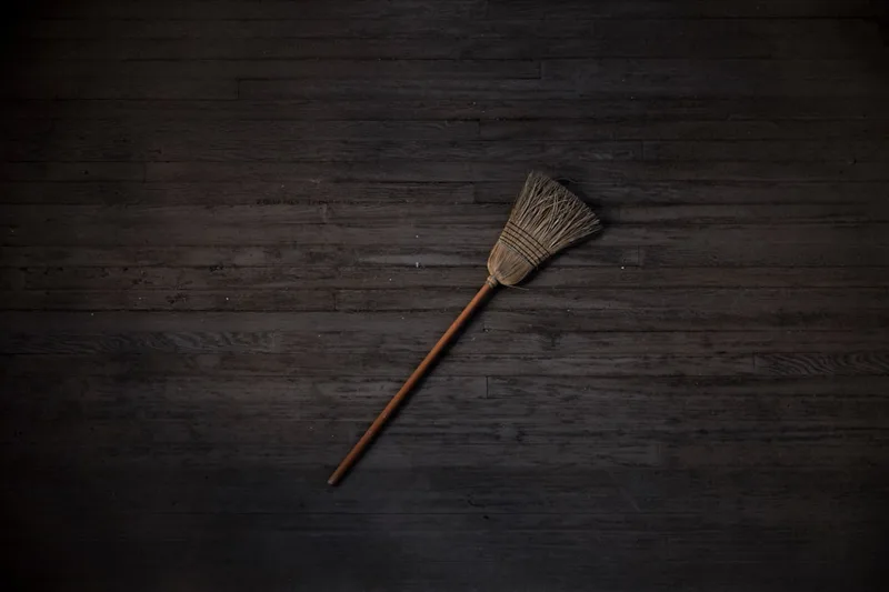 A straw broom lying across dark wood plank flooring, photographed from above in low light