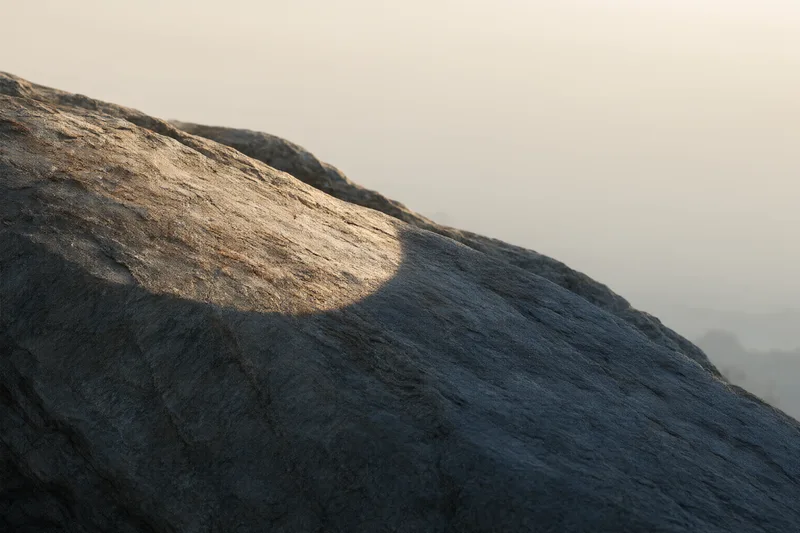 A weathered rock slope in low sunlight, a warm patch of light on its upper face while the rest of the surface sits in cool blue-grey shadow, pale mist behind