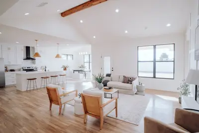 Salón y cocina abiertos y luminosos, con suelo de madera clara, sofá blanco, sillones de madera y isla de cocina con taburetes