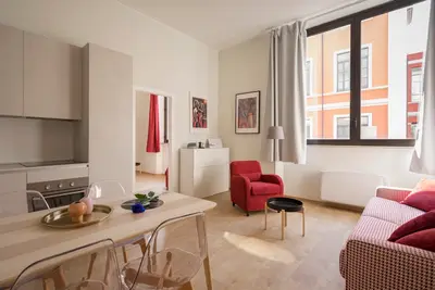A bright, minimalist studio apartment with a neatly made bed, a small desk by the window, and a plant on the windowsill, morning light streaming in
