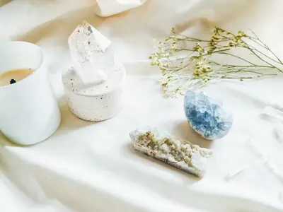 Vela blanca apagada junto a una punta de cristal de cuarzo, un cúmulo de cristal azul, una lámina de geoda y flores secas sobre tela crema