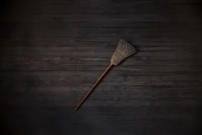 Una escoba de paja extendida sobre un suelo de tablones de madera oscura, fotografiada desde arriba con poca luz